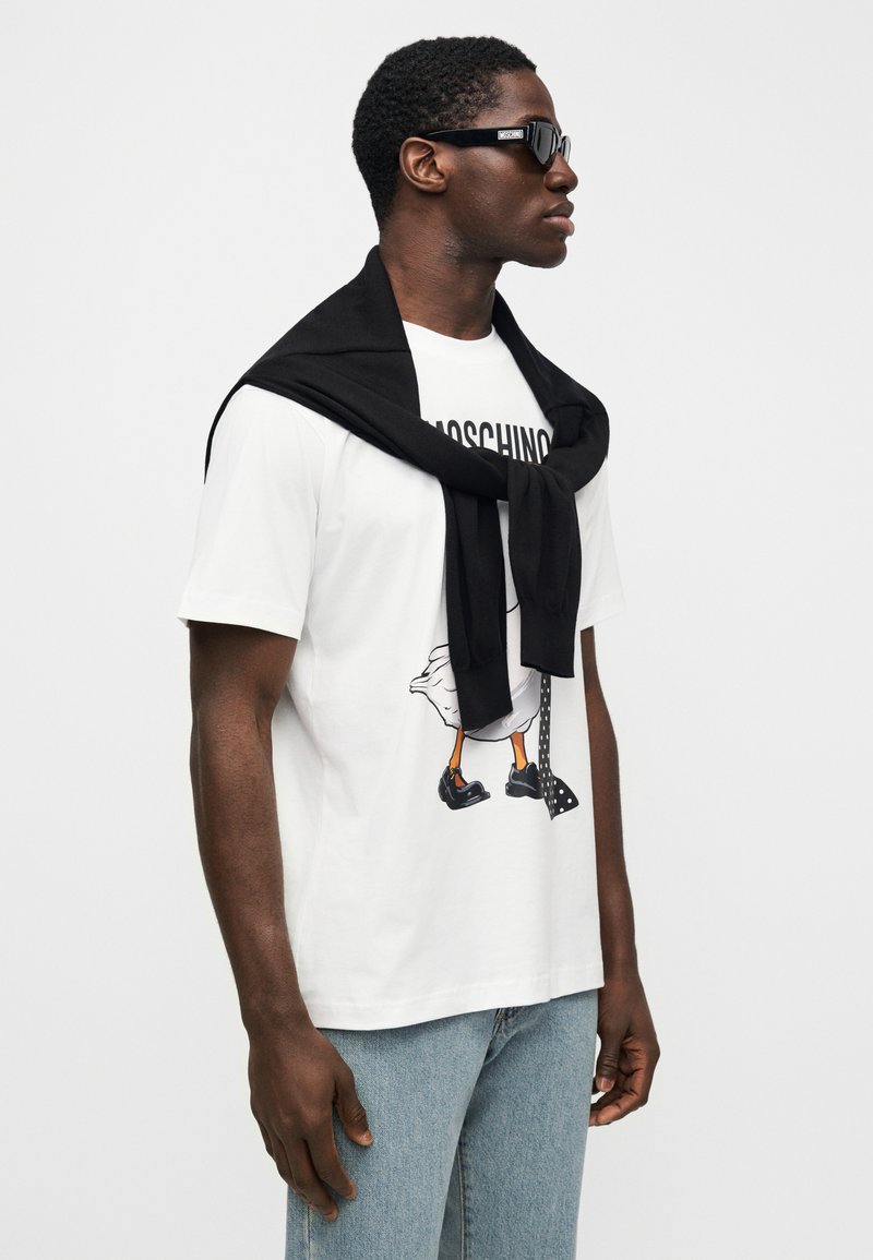 Weißes T-Shirt mit einem Cartoon-Design, das eine Ente und eine Tupfenkrawatte zeigt, kombiniert mit hellblauen Jeans und einem schwarzen Pullover, der um den Hals gebunden ist.