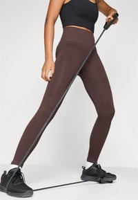 Barna csíkozott leggings, magas derékkal, fekete crop top-pal és fekete sportcipővel párosítva, amelyben egy ellenállás szalagot tart.