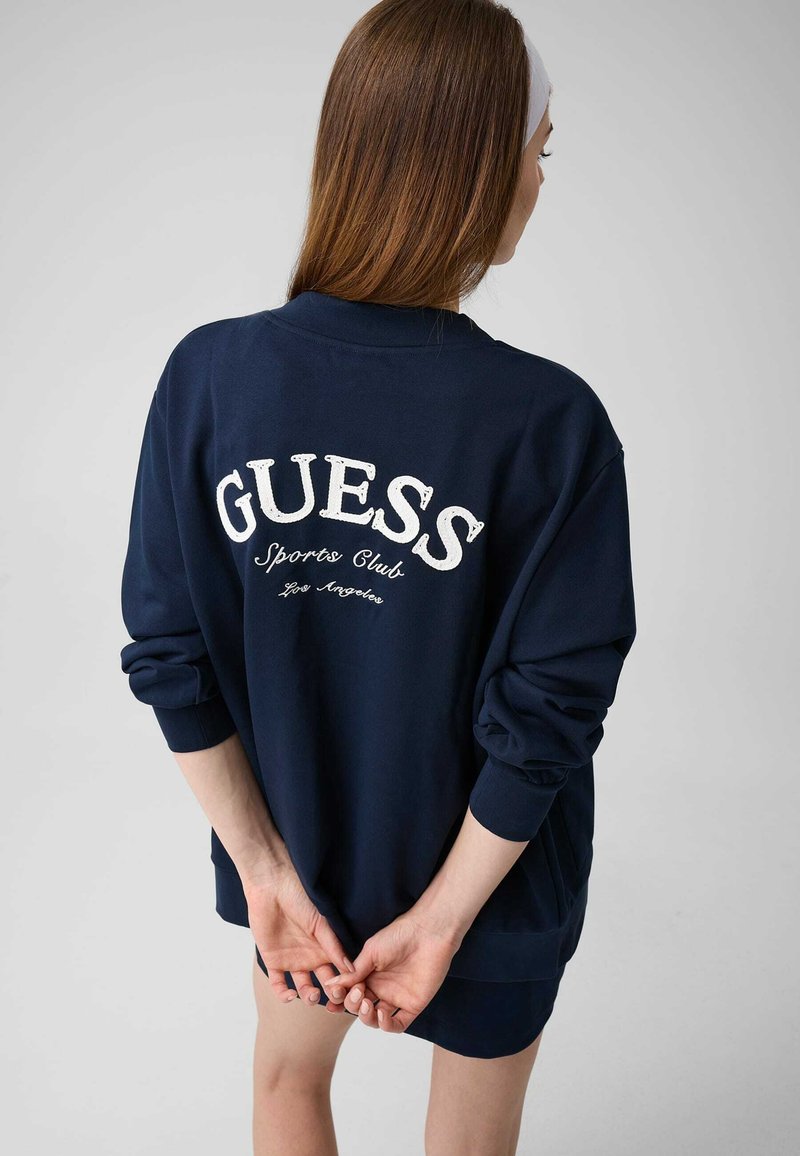 Femme aux longs cheveux bruns et bandeau blanc portant un sweat-shirt bleu marine avec le texte "GUESS Sports Club Los Angeles" dans le dos.