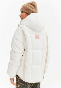 Biała pikowana kurtka z fakturowanym wykończeniem, z różową naszywką, beżowymi akcentami i dużym kapturem. W połączeniu z spodniami w panterkę.