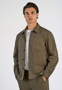 Oliven grøn jakke lavet af glat stof, med krave, knaplukning, en brystlomme og sidelommer. Hvid t-shirt under.