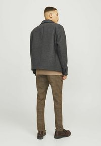 Veste à carreaux gris avec une coupe décontractée, associée à un pantalon à carreaux marron. Chaussures en cuir, texture du tissu visible sur les vêtements.