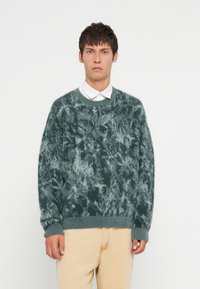 Pūkains zaļš džemperis ar teksturētu, marmora rakstu, apaļu apkakli un ribinātām piedurknēm. Kombinēts ar gaišiem džinsa biksēm.