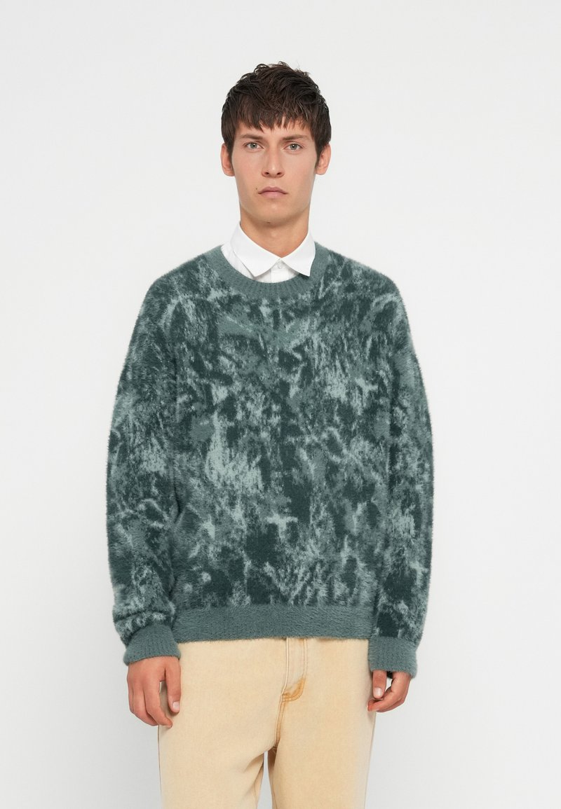 Pūkains zaļš džemperis ar teksturētu, marmora rakstu, apaļu apkakli un ribinātām piedurknēm. Kombinēts ar gaišiem džinsa biksēm.