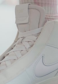 Hvit Nike høy-top sneaker med en glatt lær overdel, teksturerte detaljer, flate snørebånd og en subtil logo på tungen.