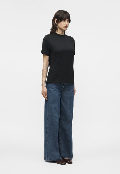 Γυναίκα που στέκεται κοιτώντας ελαφρώς προς τα δεξιά, φορώντας μαύρο τοπ με κοντά μανίκια, μπλε τζιν με φαρδιά μπατζάκια και καφέ παπούτσια σε απλό λευκό φόντο.