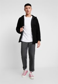 Sort fleecejakke med en sort lynlås og en lomme med klap, hvid t-shirt, grå bukser og lyserøde sneakers. Simpelt design og afslappet pasform.