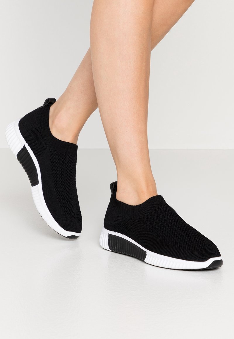 Jambes portant des baskets slip-on en tricot noir avec des semelles blanches et noires texturées, positionnées comme si elles marchaient sur un sol clair.