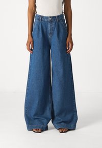 Džínové široké kalhoty v středně modré barvě, s vysokým pasem, velkými předními kapsami a volným střihem, který splývá až na zem.