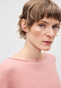 Camisola de malha rosa com textura suave, apresentando um decote largo e detalhes de costura mínimos. Modelo com cabelo curto e maquilhagem neutra.