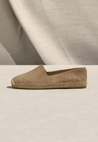 Μπεζ σουέντ εσπαντρίγια με ραμμένη σόλα από τζούτ, επίπεδη φτέρνα και σχεδιασμό slip-on, παρουσίαση σε ουδέτερο φόντο με υφασμάτινο υπόβαθρο.