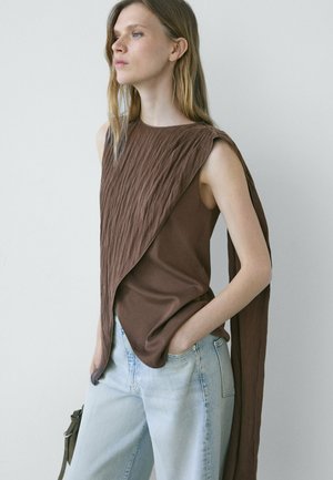 Femme aux longs cheveux blonds portant un haut marron sans manches à texture drapée et un jean bleu clair, debout avec une main dans la poche.