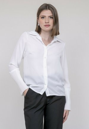 Chemise blanche à manches longues avec des poignets plissés texturés, un col classique et une finition lisse. Associée à un pantalon noir.