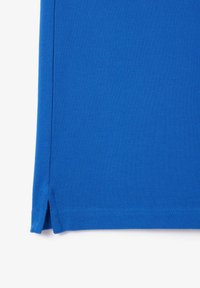 Polo azul con tela texturada, cortas aberturas laterales y dobladillo cosido. El color es vibrante y está distribuido de manera uniforme por toda la tela.