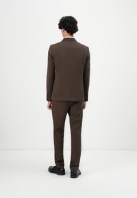 Férfi, aki háttal áll, barna zakót és hozzá illő nadrágot visel, fekete elegáns cipővel, egy sima fehér háttér előtt.