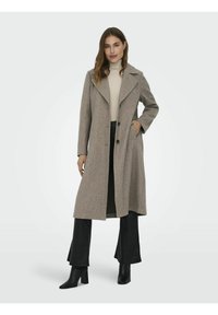 Manteau en laine beige avec des revers larges, fermeture à double boutonnage, poches latérales et coupe ajustée, associé à un pantalon noir et des bottes à talons.