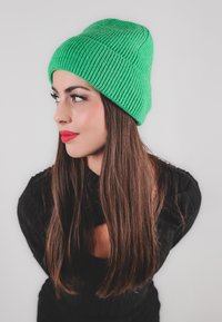 Cappello a cuffia in maglia verde brillante con un design a coste testurizzate, indossato leggermente inclinato. Il modello ha i capelli lunghi castani ed è vestito con una maglietta nera.