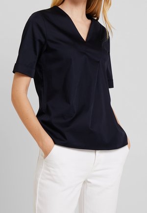 Vrouw met een zwart V-hals shirt met korte mouwen en omgeslagen manchetten, en witte broek, handen in de zakken tegen een effen achtergrond.