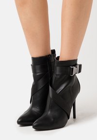 Botines negros de cuero texturizado, con puntera puntiaguda, tacón alto de aguja y correas cruzadas con un detalle de hebilla plateada.