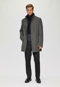 Grau melierter Wollmantel mit strukturiertem Finish, Stehkragen, Reißverschlusstaschen und Knopfdruck, kombiniert mit schwarzen Hosen und Schuhen.