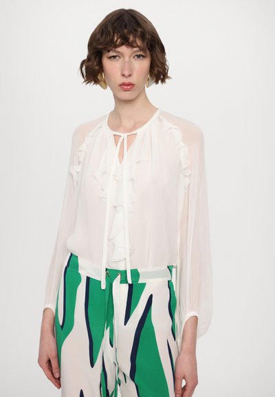 Blouse blanche avec détail volanté à l'avant et col noué. Associée à un pantalon vert et blanc à motifs. Texture de tissu lisse et design fluide.
