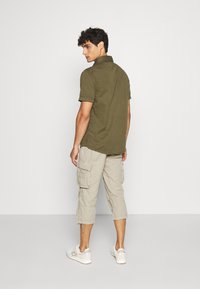 Chemise à manches courtes vert olive, pantalons cargo beige clair avec poches latérales, baskets blanches, mannequin de dos devant un fond neutre.