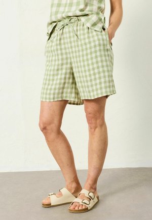 Person trägt hellgrüne und weiße karierte Shorts und passendes Oberteil, steht barfuß auf grauem Boden vor weißer Wand.