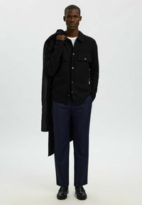 Giacca camicia nera in lana con due tasche frontali e chiusura a bottoni, indossata sopra una camicia bianca. Abbinata a pantaloni blu navy e scarpe nere.