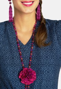 Bluse mit Textur in Blau und V-Ausschnitt, ergänzt durch eine lange Perlenkette mit roten und pinken Perlen sowie einem großen Akzent in Form einer pinken Blume.