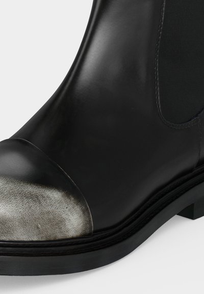 Gros plan d'une botte noire en cuir à la cheville avec un embout métallique argenté usé et un panneau latéral élastique.