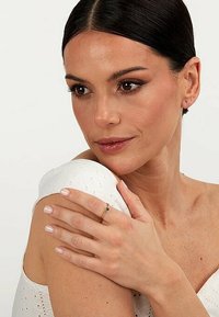 Guldring med en liten grön ädelsten, bärs på ett finger. Modellen har en ljus hy och är klädd i en vit topp med hålmönster.