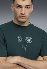 Camiseta de algodón verde oscuro con logotipos impresos, cuello redondo y mangas cortas. Incluye el logotipo de Puma y el emblema del Manchester City.