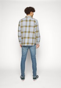 Camisa de botones en patrón de cuadros azules y olivas; mangas largas, cuello estándar, combinada con jeans de color azul claro y zapatillas negras y blancas.