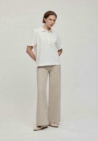 Polo shirt bianco a maniche corte con patta di bottoni, abbinato a pantaloni a palazzo beige chiaro. Il modello si trova su uno sfondo neutro.