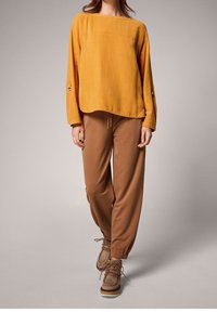 Maglietta gialla senape a vestibilità morbida con scollatura ampia e maniche lunghe, abbinata a pantaloni marroni a sigaretta e scarpe marroni con lacci.