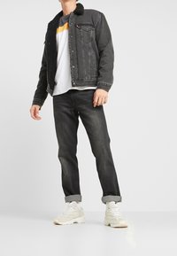 Henkilö, jolla on päällään musta sherpa-vuorattu denimhousujakku, valko- ja keltaraidallinen paita, mustat käärityt farkut ja valkoiset lenkkarit, seisoo.