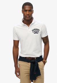 Camisa polo blanca con logotipo azul marino, de manga corta y con cuello. Pantalones beige con un cinturón oscuro y un suéter azul marino atado a la cintura.