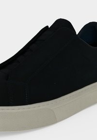 Μαύρο παπούτσι τύπου sneaker χαμηλού ύψους με λευκή υφή στη σόλα από καουτσούκ και ελαστικά κορδόνια, εμφανίζεται από κοντινή πλευρική γωνία σε λευκό φόντο.