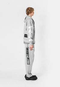 Sudadera gris con detalles gráficos en blanco, combinada con pantalones grises que presentan etiquetas impresas, llevada con zapatos negros sin cordones.