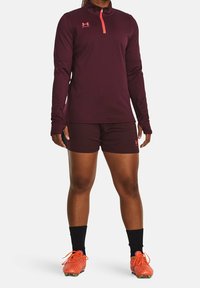 Tummanpunainen pitkähihaisen paita, jossa on neljänneksen vetoketju ja vaaleanpunaiset yksityiskohdat, yhdistetty tummanpunaisiin shortseihin ja oransseihin jalkapallokenttäkengiiin, mustat polven yläpuolelle ulottuvat sukat.