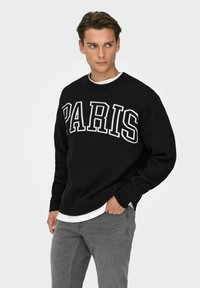 Pull en maille noire avec le mot "PARIS" en lettres blanches épaisses. Col et poignets côtelés. Porté sur une chemise blanche, associé à un jean gris.