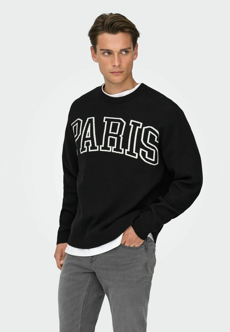 Pull en maille noire avec le mot "PARIS" en lettres blanches épaisses. Col et poignets côtelés. Porté sur une chemise blanche, associé à un jean gris.