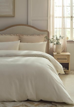 Bett mit beigem gepolstertem Kopfteil, weißen und beigen Kissen, weißer Bettdecke, hölzernem Nachttisch mit Pflanzenvase neben Fenster mit transparenten Vorhängen.