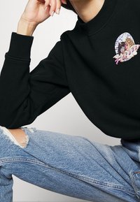 Sudadera negra con un gráfico bordado redondo que presenta texto en rosa, combinada con jeans de mezclilla azul desgastados. Puños y escote acanalados.