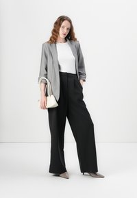 Šedý blazer, bílý tričko, černé široké kalhoty, držící malou bílou kabelku, béžové lodičky s špičatou špičkou, hladké materiály, minimalistický design.