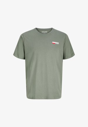 Garçon debout portant un t-shirt vert à manches courtes, un short cargo beige, des chaussettes blanches et des baskets blanches sur fond uni.