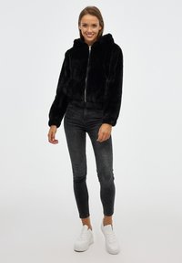 Chaqueta con capucha de piel sintética negra, con cierre mediante cremallera en la parte delantera, dobladillo elástico y mangas largas, combinada con jeans ajustados oscuros y zapatillas blancas.