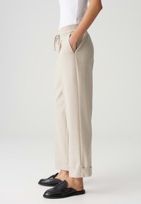 Beige, ruime broek met een elastische tailleband, zijzakken en omgeslagen boorden; gedragen met zwarte instap loafers. Soepele stof.