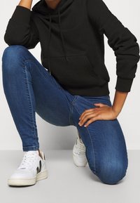 Sudadera negra con bolsillo canguro, vaqueros slim fit azules y zapatillas blancas con detalles negros, enfocándose en líneas limpias y un estilo casual.