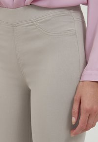 Beige elastické kalhoty mají hladký povrch, jemný design kapes a zesílené švy; jsou kombinovány s lehkým růžovým topem s dlouhým rukávem.